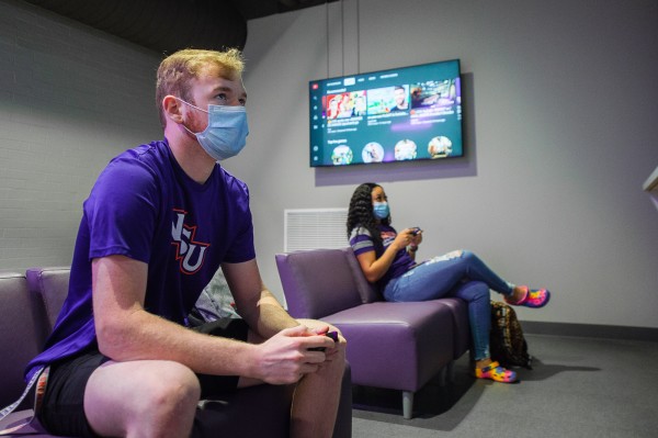 A masked male and female student sit holding game controllers while facing an off-camera screen.