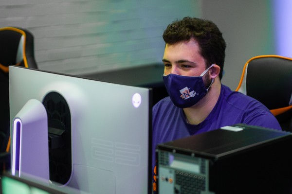 A masked male student sits at a desktop PC with an alienware monitor.