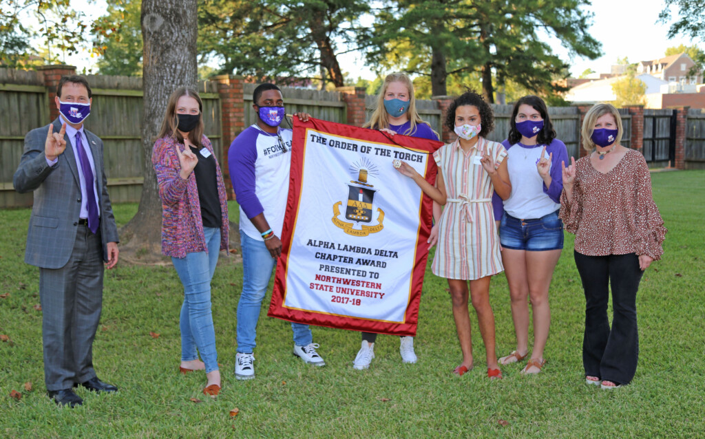 Five students in a grassy area hold up the Order of the Torch banner. Chris Maggio stands on the left, and Reatha Cox stands on the right.