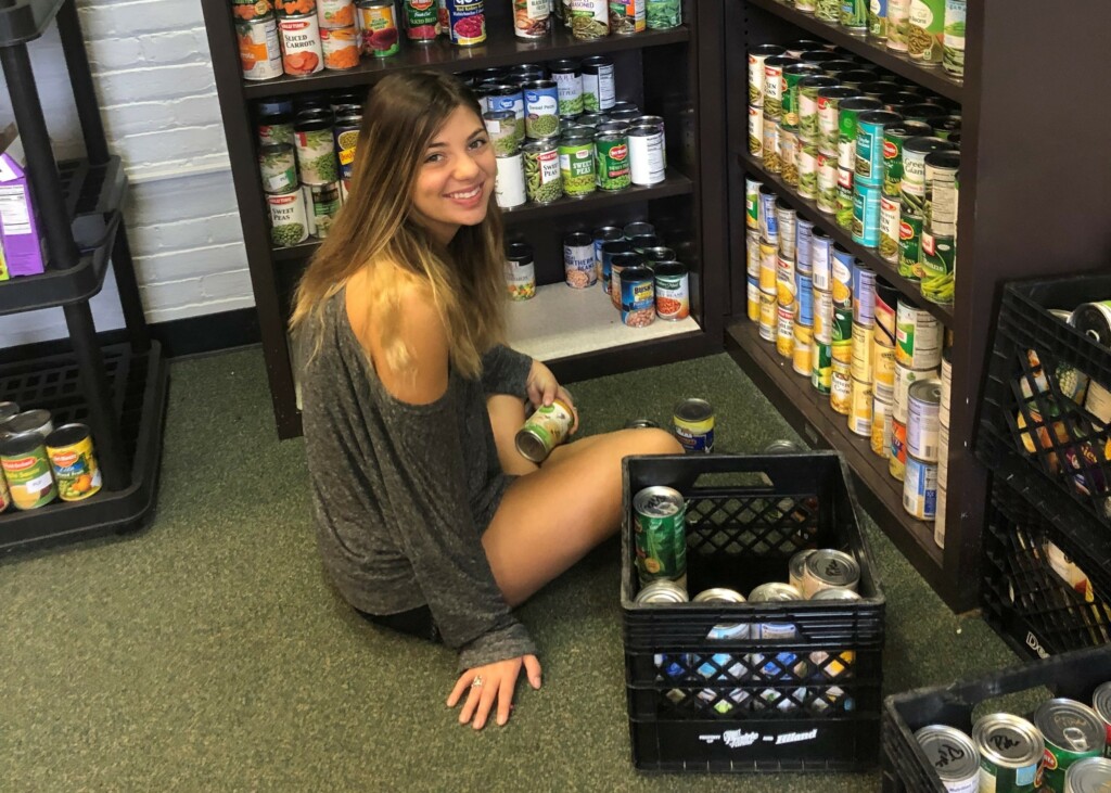 A smiling young woman places cans on a shelf while sitting on the floor.
