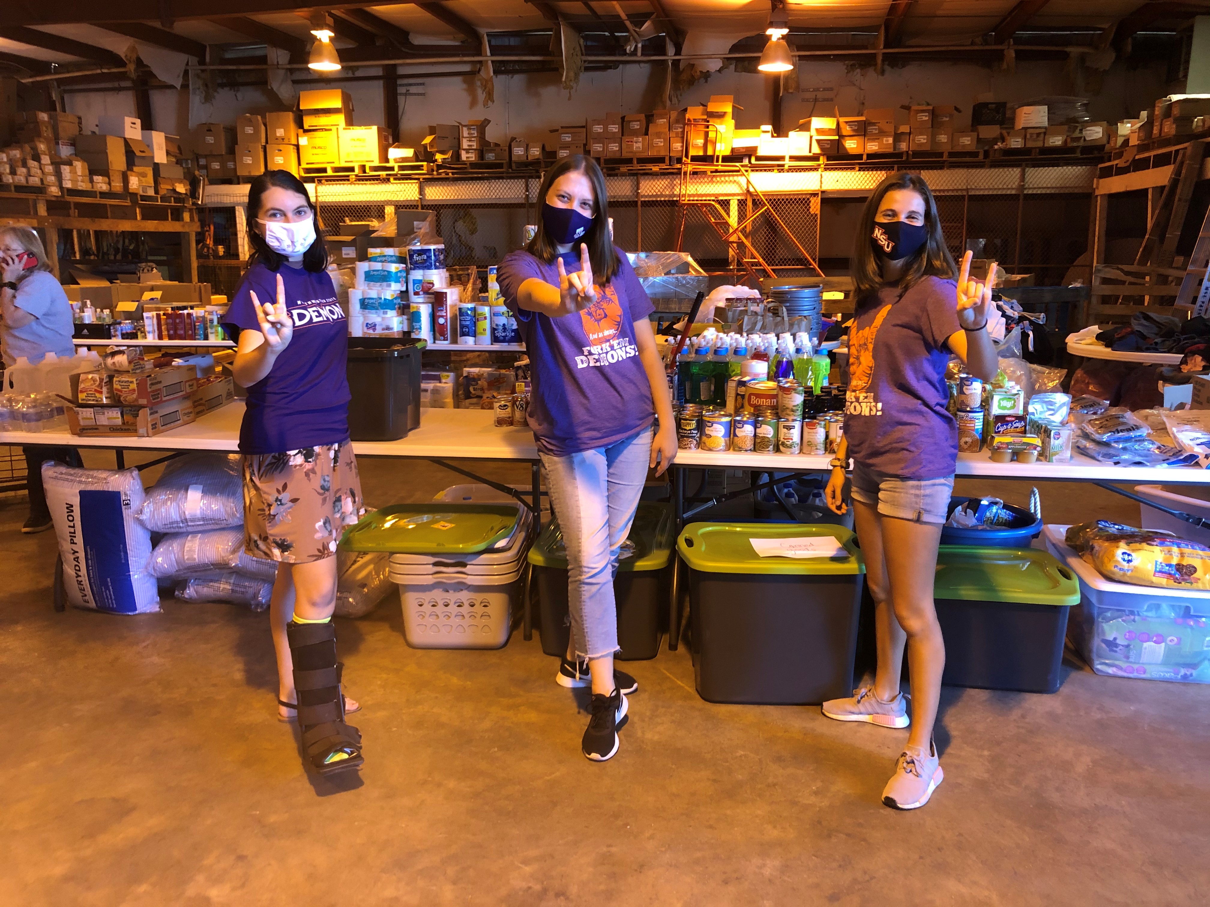 Three students pose in a warehouse in front of tables stacked with food, toiletries, and other hurricane supplies.
