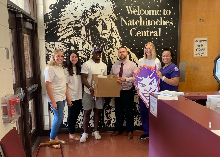 Students hold a cardboard box and NSU Demon cutout in front of the Welcome to Natchitoches Central mural.