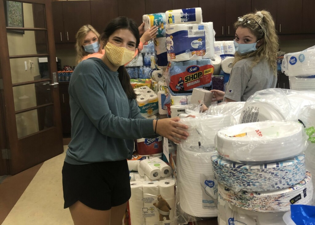 Three mask-wearing students arrange paper towels, toilet paper, and disposable plates.