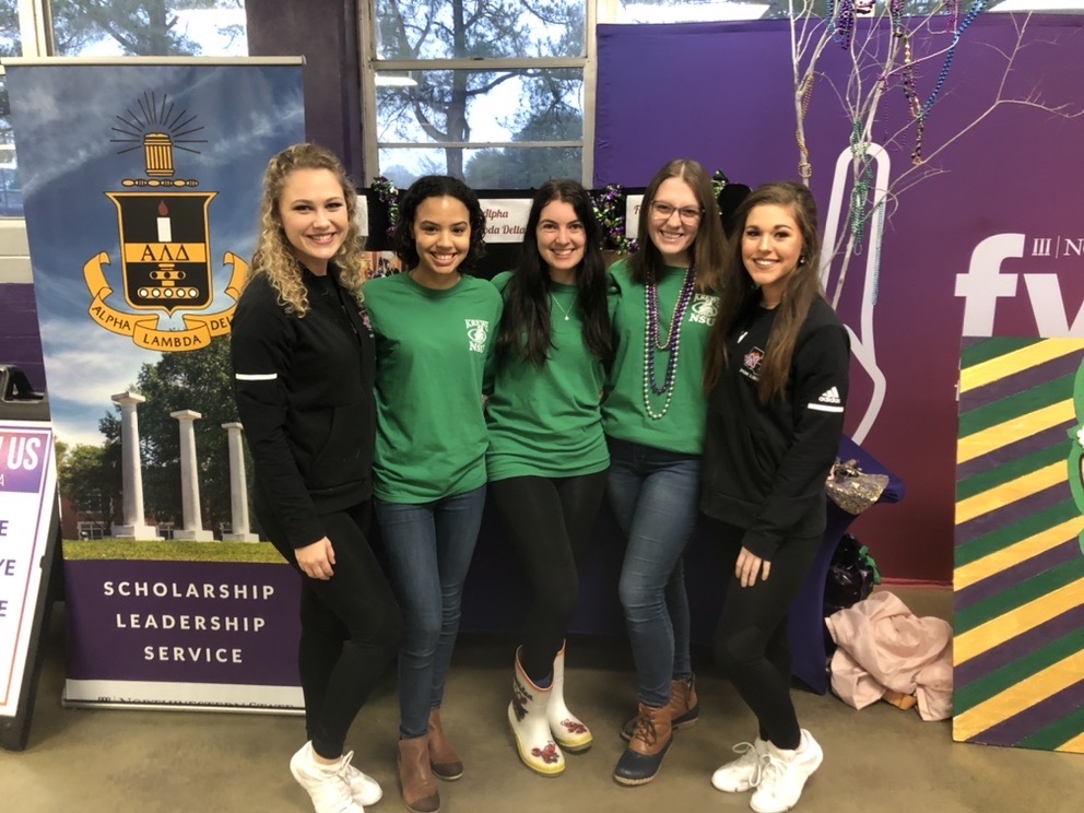 Five smiling students pose surrounded by Mardi Gras decorations and an Alpha Delta Lambda banner.