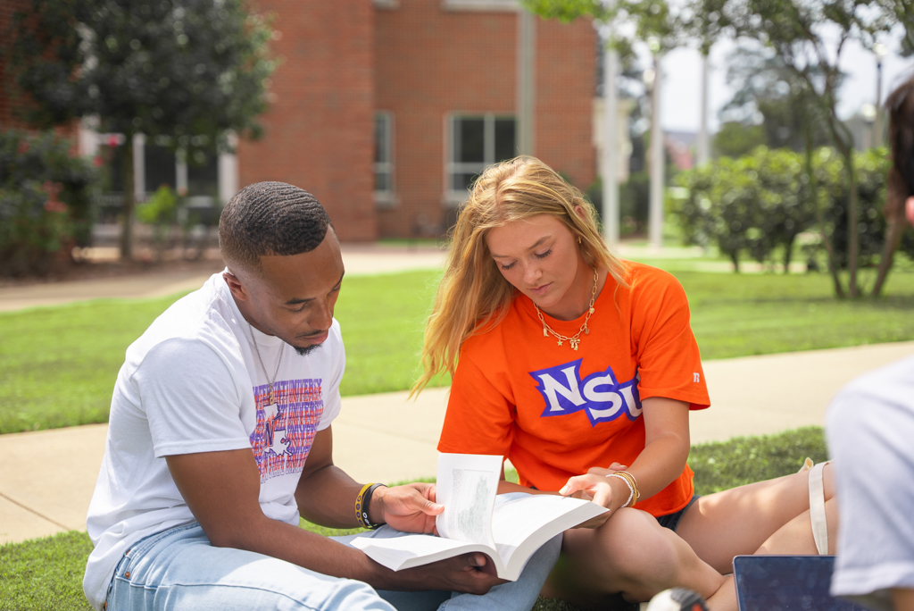 2 NSU students reading a book on the Caspari Hall lawn