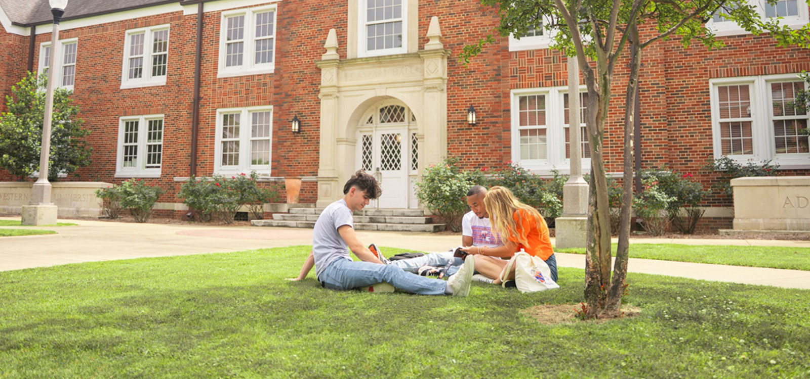 Three NSU student sitting on lawn in front of Caspari Hall.
