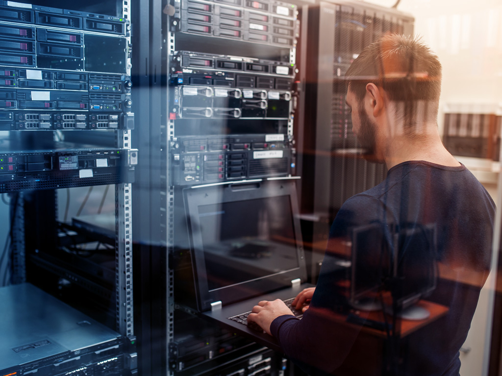 A person is standing in a server room, working on a laptop connected to a server rack filled with network equipment.