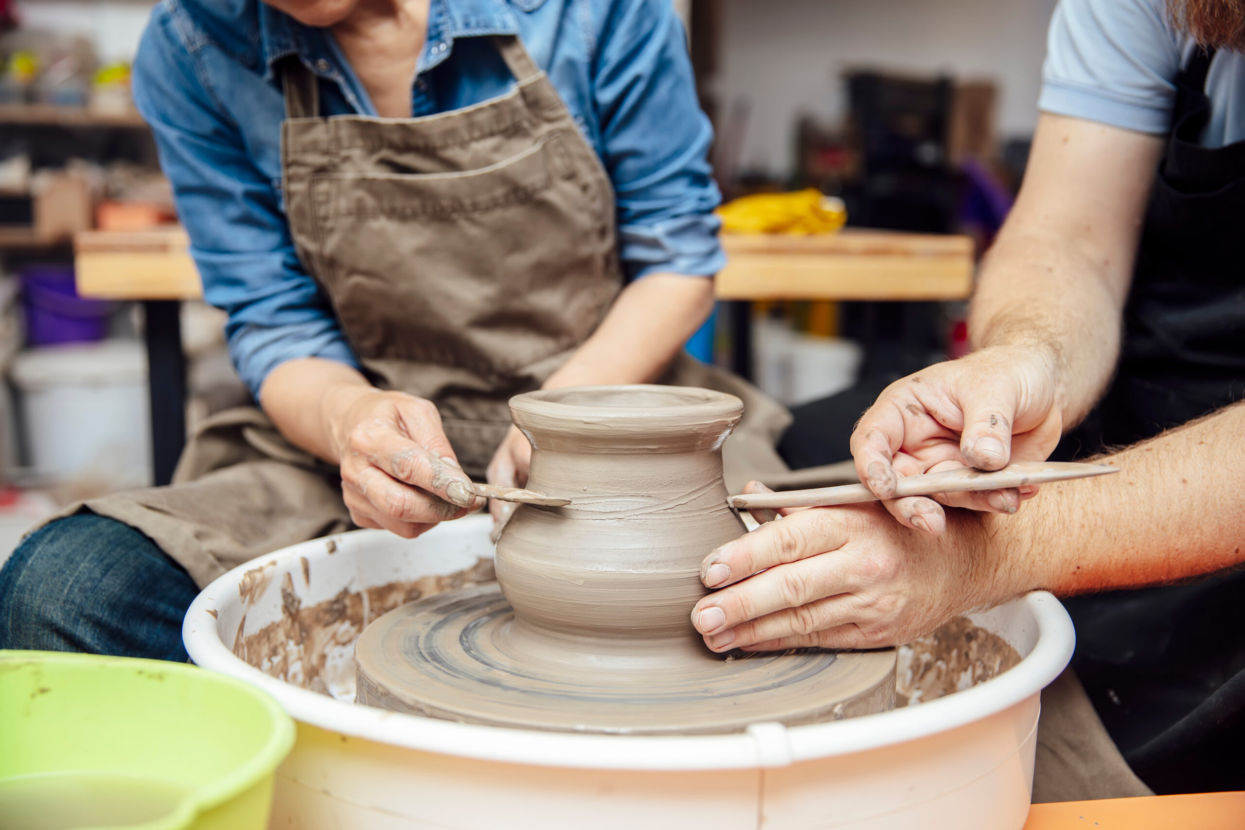 Senior woman spinning clay on a wheel with a help of a teacher at pottery class