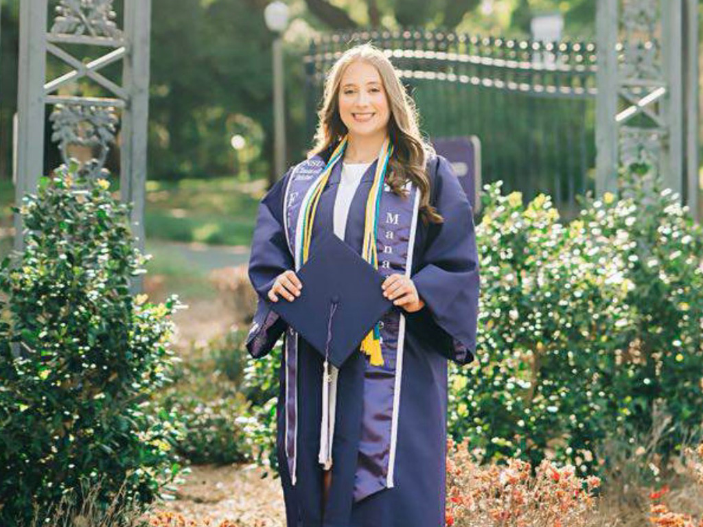 A woman wearing a graduation gown and honor cords stands outdoors, holding a graduation cap, with greenery and an iron gate in the background.
