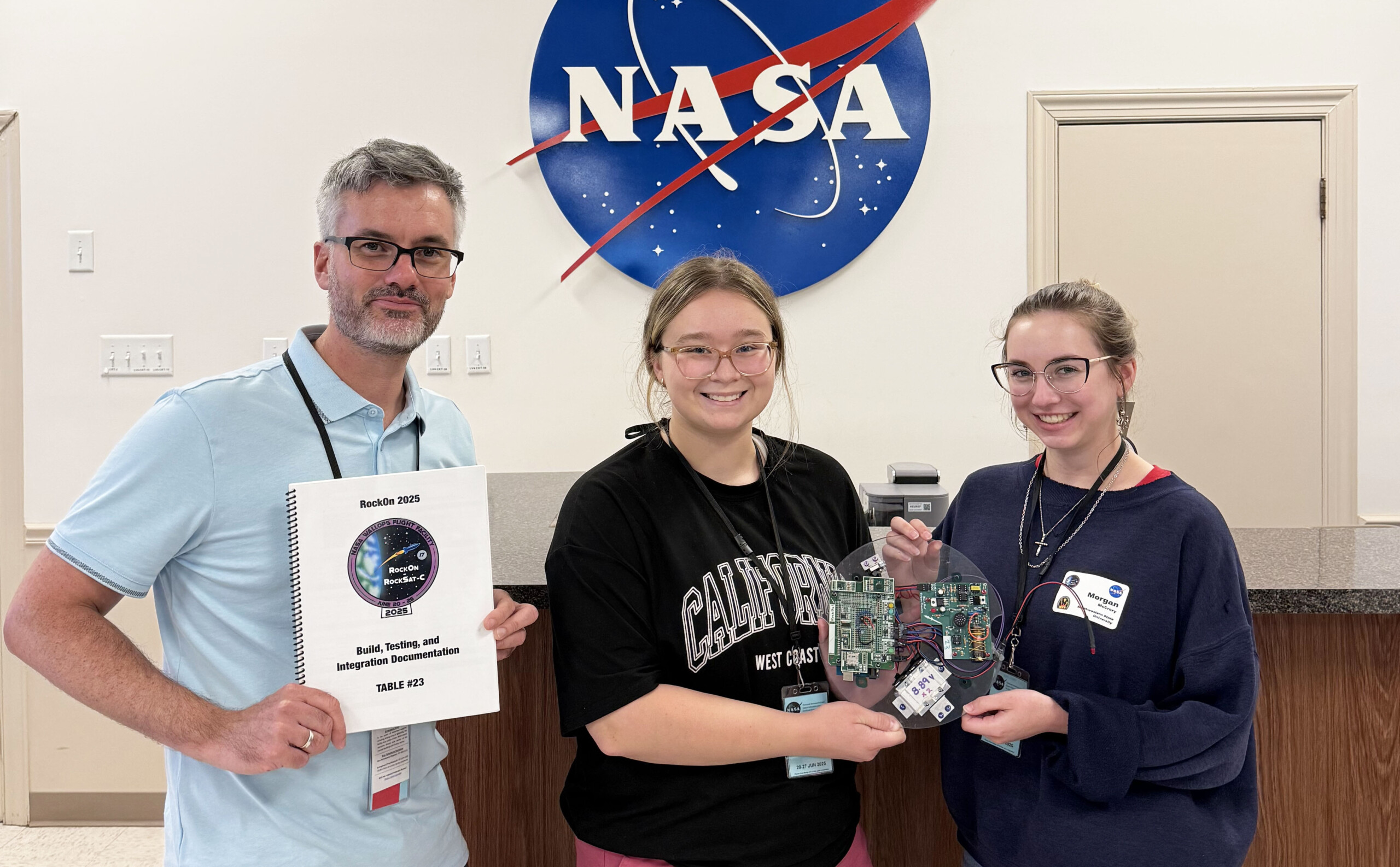 Dr. Damien Tristant, Shae Wetzel and Morgan McCrory, from left, are shown during the 2025 RockOn program at NASA’s Wallops Flight Facility on Wallops Island, Virginia, holding the checklist manual and their assembled payload after completing the soldering and coding stages.