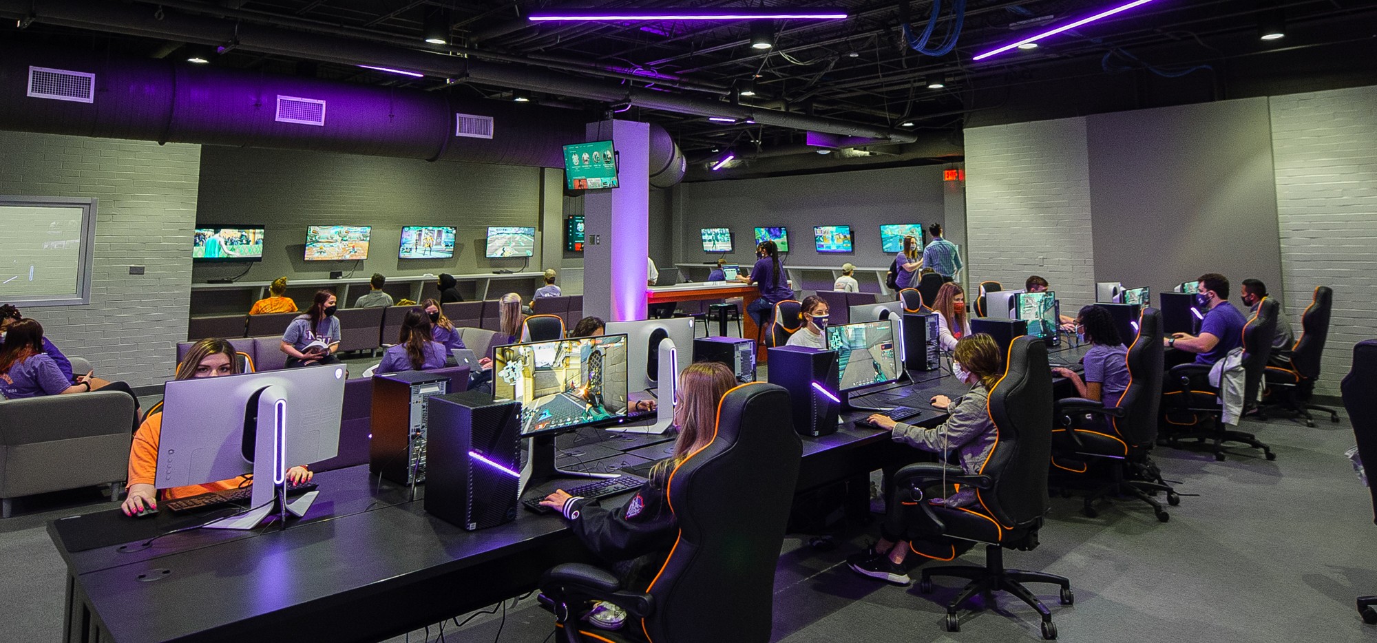 Masked students sit in a purple-lit computer lab engaging in a gaming competition.